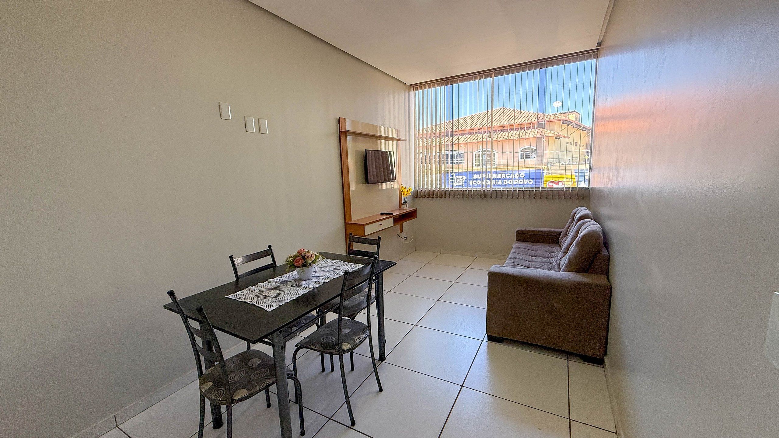 A small, well-lit room with a dining table, chairs, a sofa, and a TV mounted on the wall.