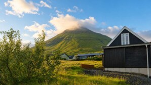 Een landelijk landschap met een berg op de achtergrond, een houten gebouw met een puntige dakschildering, en een grasachtige voorgrond.