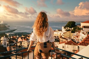 Eine Person steht auf einem Balkon mit Blick auf eine Küstenstadt, mit Gebäuden und einem Kreuzfahrtschiff in der Ferne.