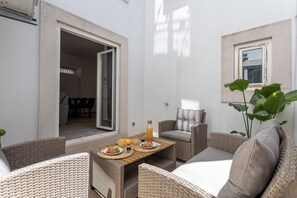 A sunlit patio with wicker furniture, a table set with plates and drinks, and a view into the adjacent room. - Baixa Garden: 2BR + Patio | Perfect for Families (Lisboa)