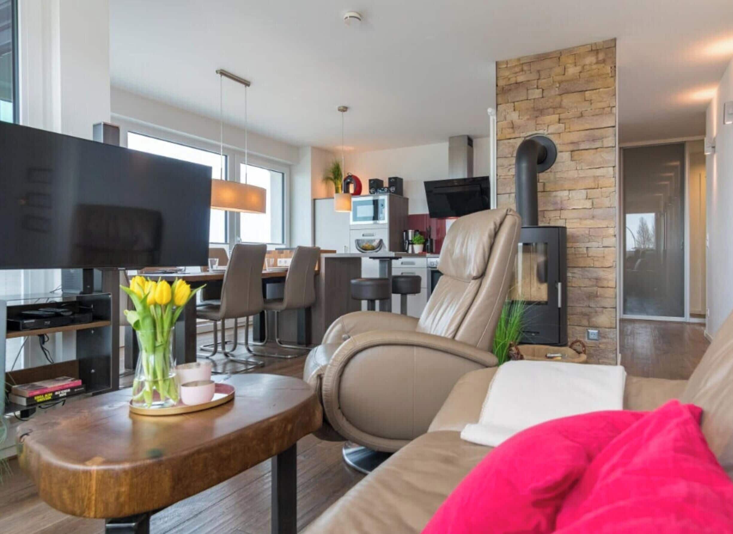 A modern living room with a sofa, a TV, a coffee table with flowers, and a kitchen area in the background.