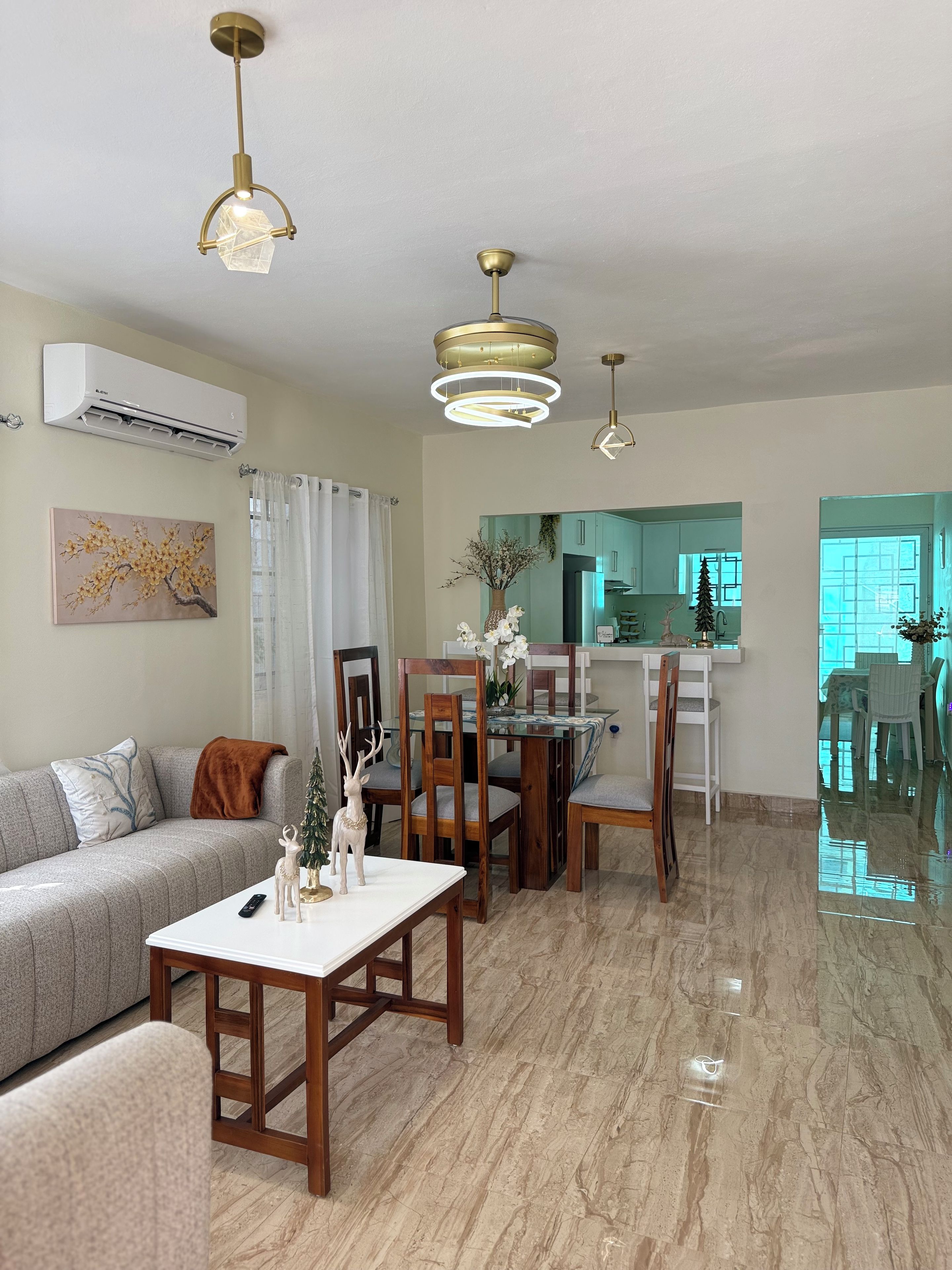 A modern living room with a sofa, dining table, and chairs, a kitchen in the background, and a ceiling with pendant lights.