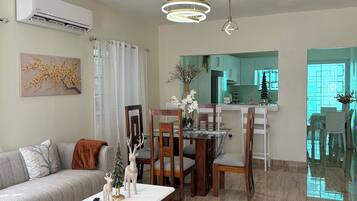 A modern living room with a sofa, dining table, and chairs, a kitchen in the background, and a ceiling with pendant lights.