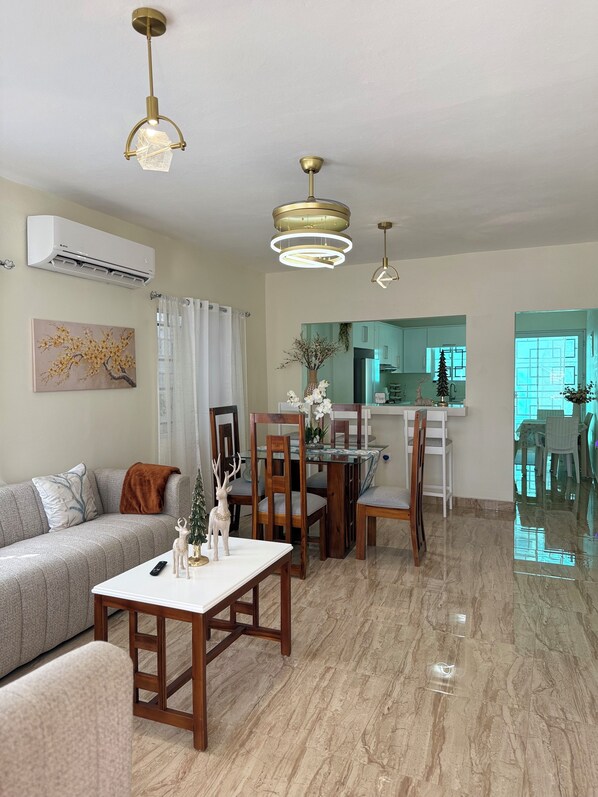 A modern living room with a sofa, dining table, and chairs, a kitchen in the background, and a ceiling with pendant lights.