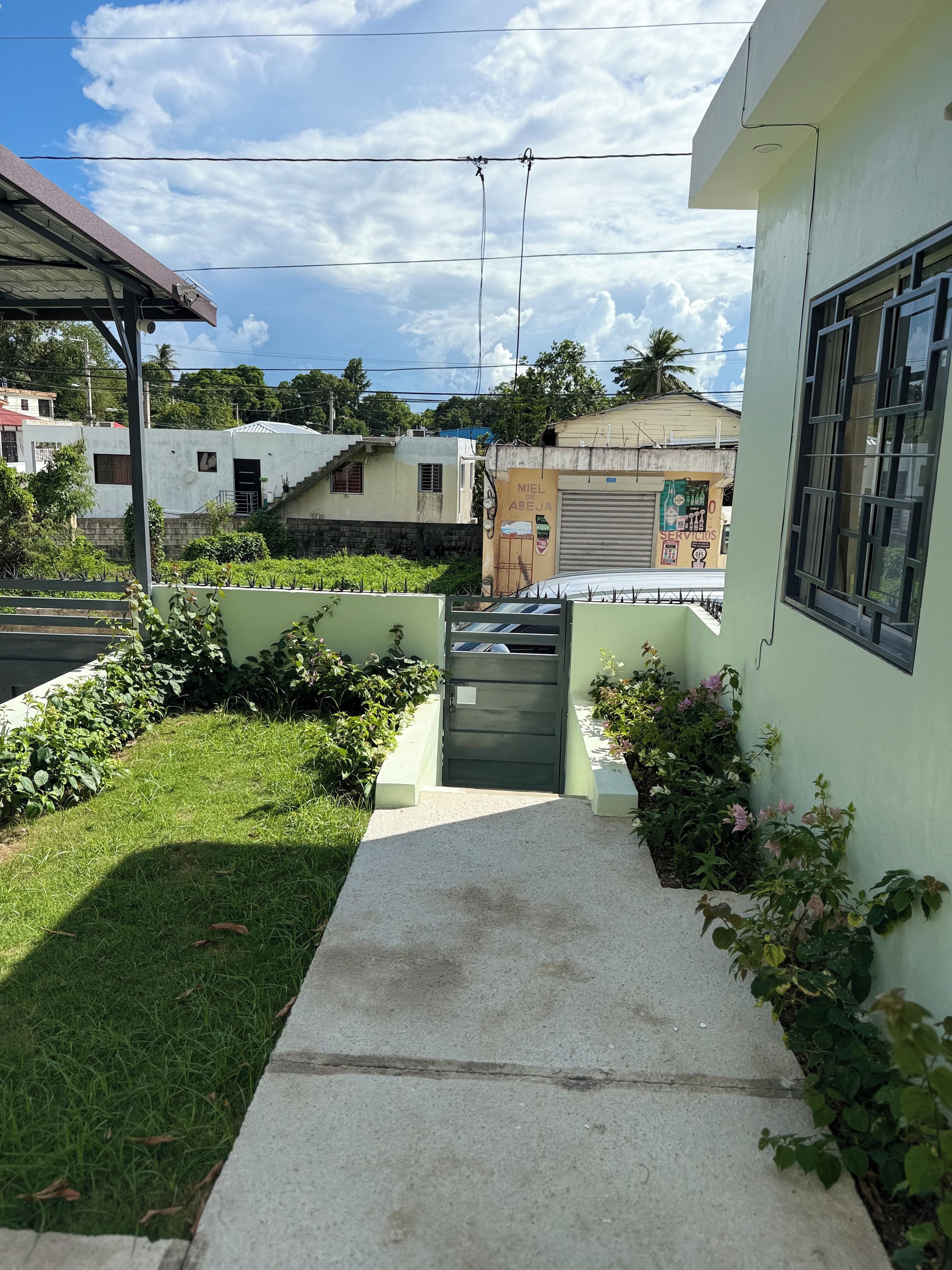 A residential area with a paved pathway leading to a gated entrance, surrounded by greenery and adjacent buildings.