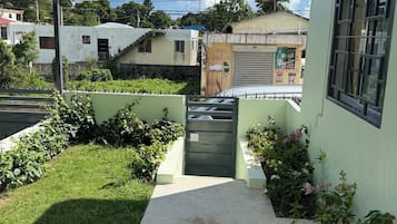 A residential area with a paved pathway leading to a gated entrance, surrounded by greenery and adjacent buildings.