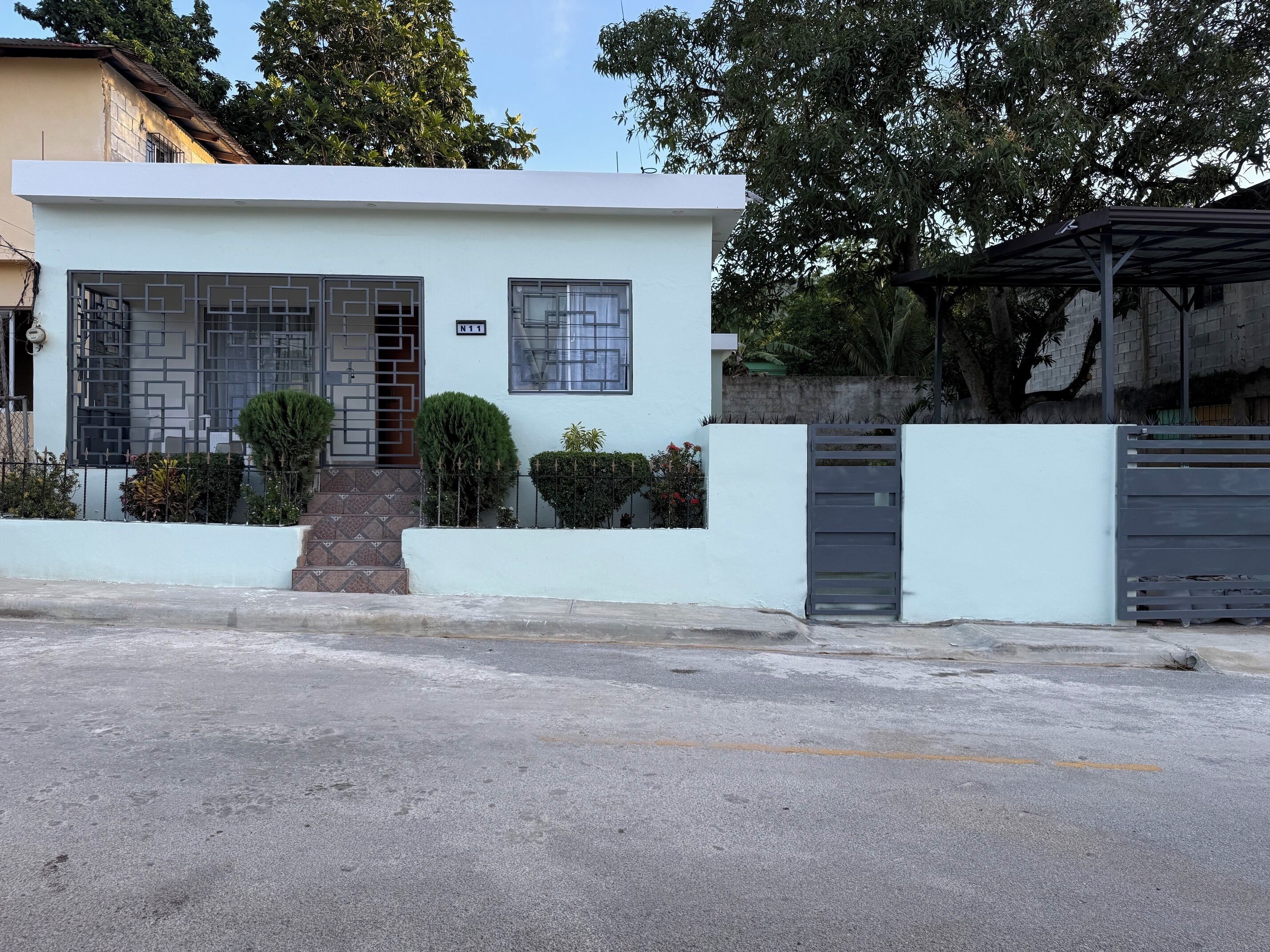 Una casa moderna de dos pisos con fachada blanca, entrada protegida por un portón y un pequeño jardín delantero con arbustos podados.