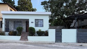 Une maison moderne à deux étages, avec une façade blanche, une entrée sécurisée par un portail et un petit jardin devant, agrémenté d’arbustes taillés.