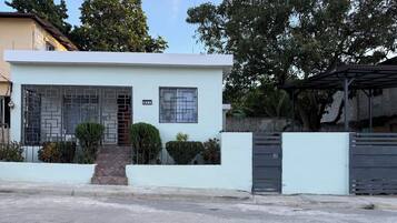 Una casa moderna de dos pisos con fachada blanca, entrada protegida por un portón y un pequeño jardín delantero con arbustos podados.