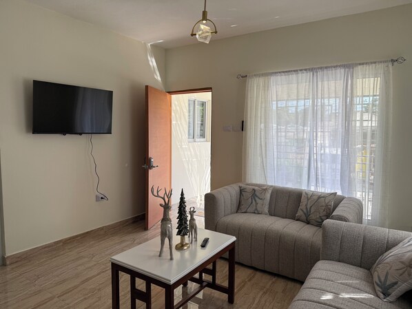 Ein Wohnzimmer mit einer Couch, einem Couchtisch, einem an der Wand befestigten Fernseher und einem Fenster mit Vorhängen.