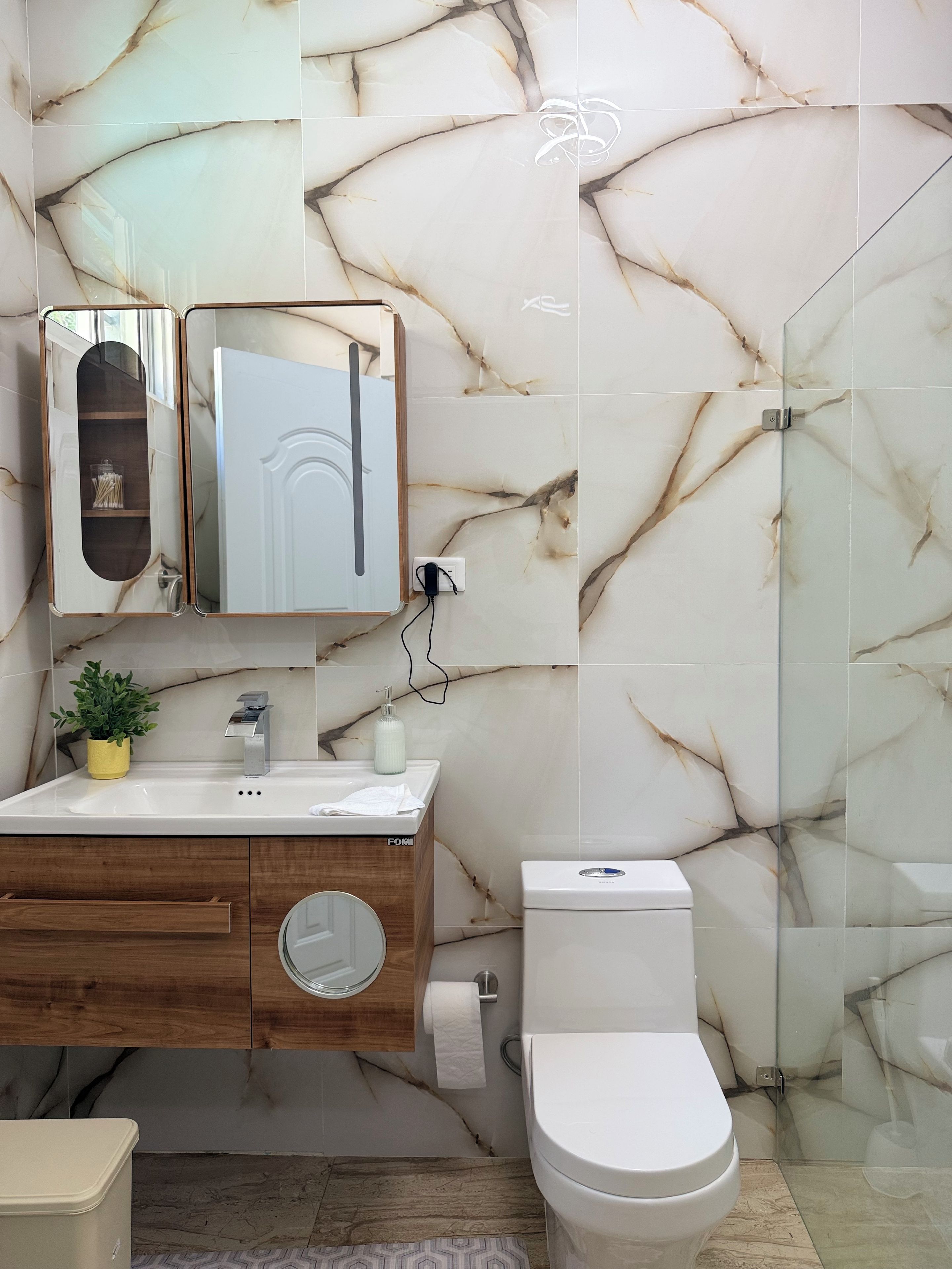 Un baño con salpicadero de mármol, tocador de madera con espejo, lavabo, inodoro y cabina de ducha con mampara de vidrio.