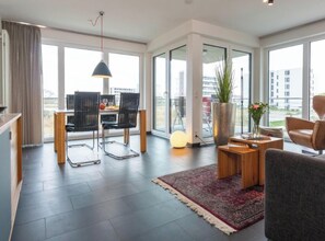 A modern living room with a dining area, large windows, a potted plant, and a round table with chairs. - Holiday Apartment With 1 Bedroom (Heiligenhafen)