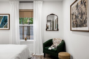 Ein Schlafzimmer mit einem Bett, einem grünen Sessel, einem Fenster mit Vorhängen und zwei gerahmten Kunstwerken.