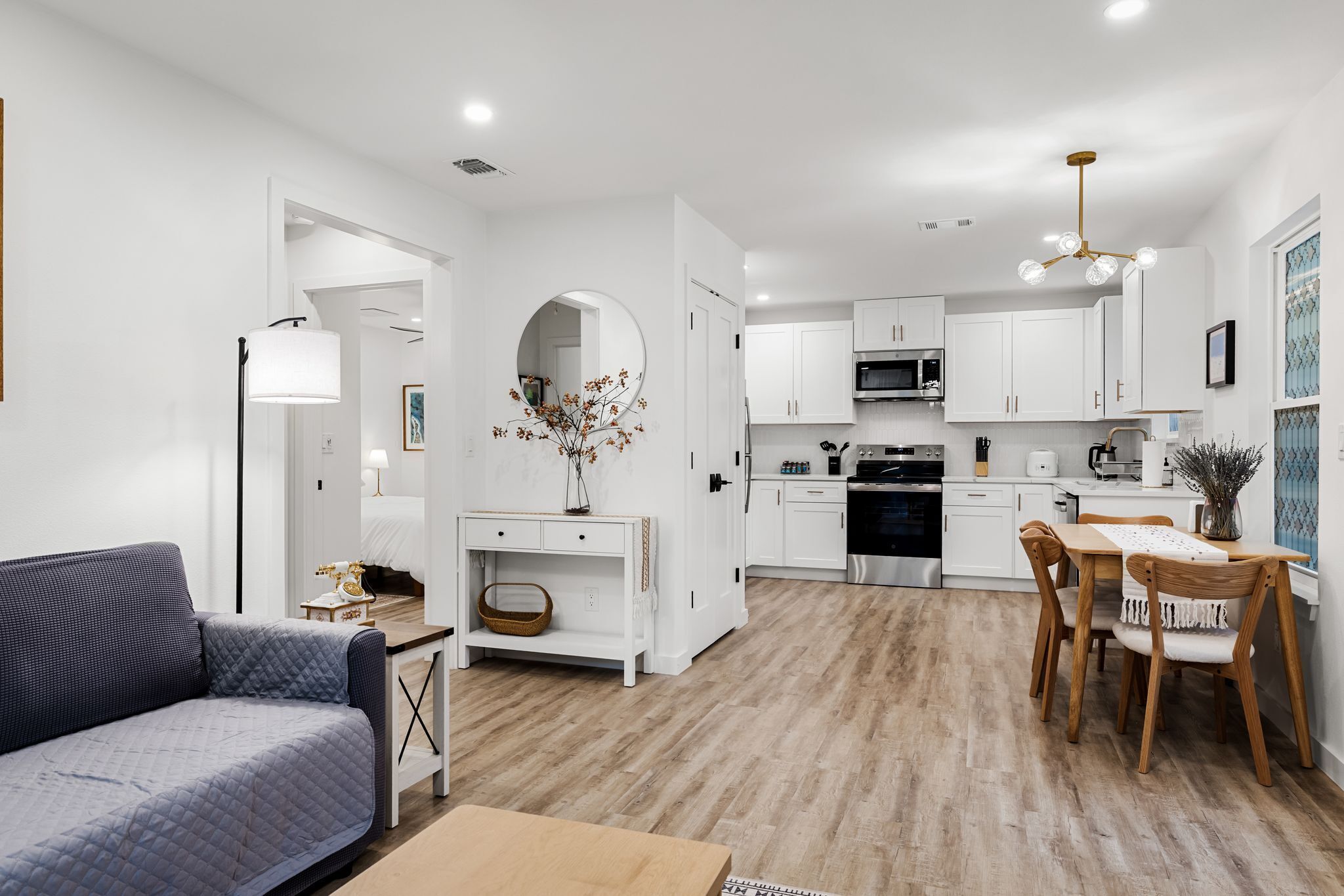 A modern kitchen with white cabinets, a dining area with a wooden table and chairs, and a living room with a sofa and a lamp.