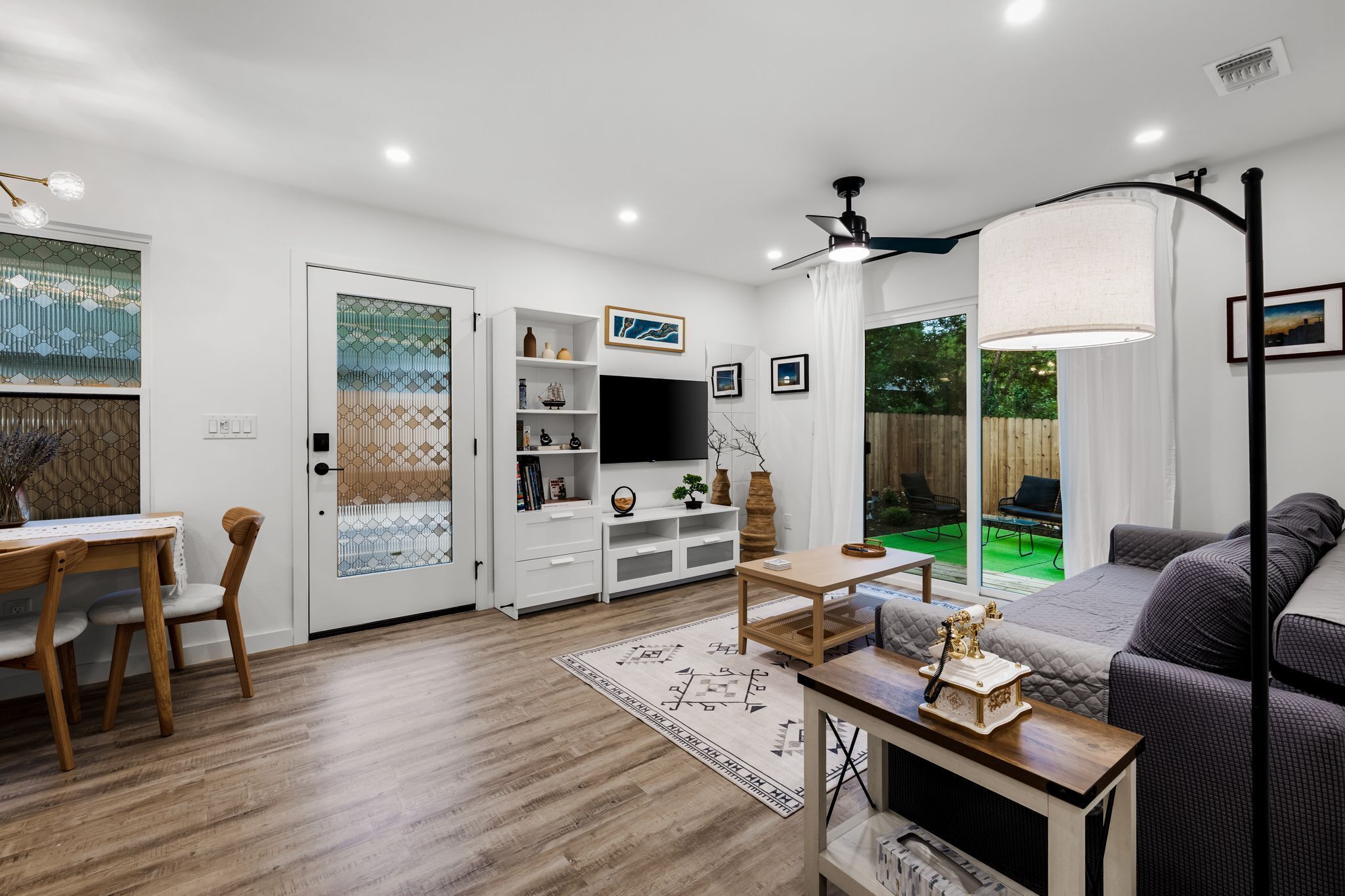 A modern living room with a flat-screen TV, a dining area with a wooden table and chairs, and a cozy seating area with a sofa and a lamp.