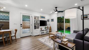 A modern living room with a flat-screen TV, a dining area with a wooden table and chairs, and a cozy seating area with a sofa and a lamp.