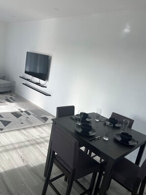 Uma sala de jantar moderna com mesa de madeira escura posta para quatro pessoas, televisão de tela plana na parede e tapete estampado no chão.