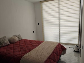 Ein Schlafzimmer mit einem Bett, Kissen, einem Fenster mit Jalousien und einer Fußbodenheizung.
