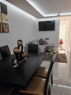 A modern living room with a flat-screen TV mounted on the wall, a grey sofa with cushions, a dining table with chairs, and a coffee table.