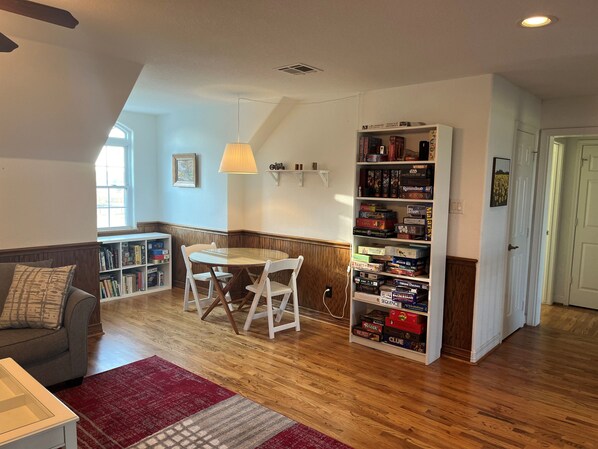 A cozy living room with a dining table, a bookshelf filled with board games, a red rug, and a grey sofa. - Large Country 7000 SF Estate On 68 Acres (Paige)
