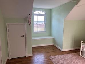 A room with a door, a window with a view, a bench, and a wooden floor.