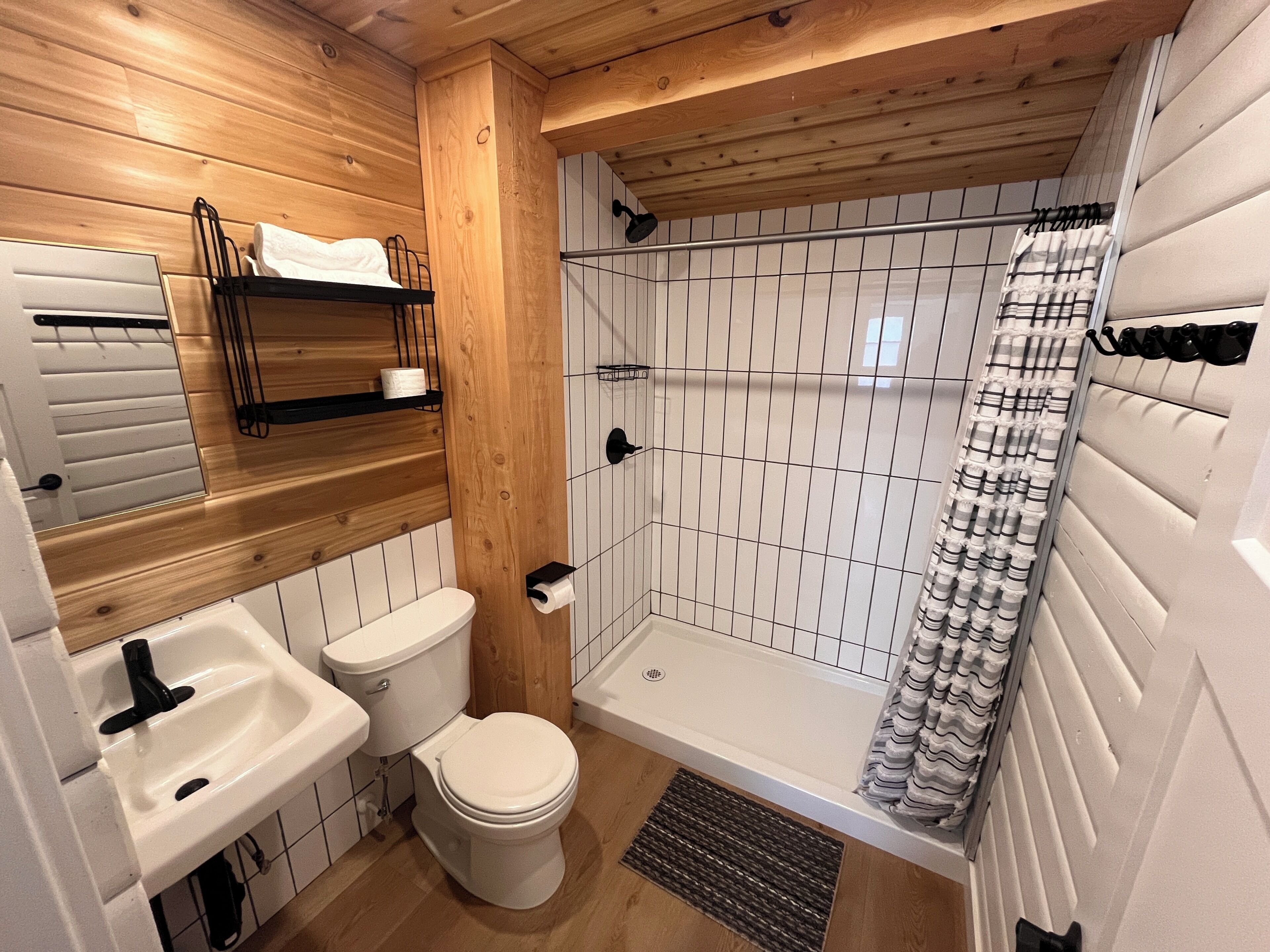 A bathroom with a wooden ceiling, white tiled walls, a white sink, a toilet, and a shower with a striped curtain.