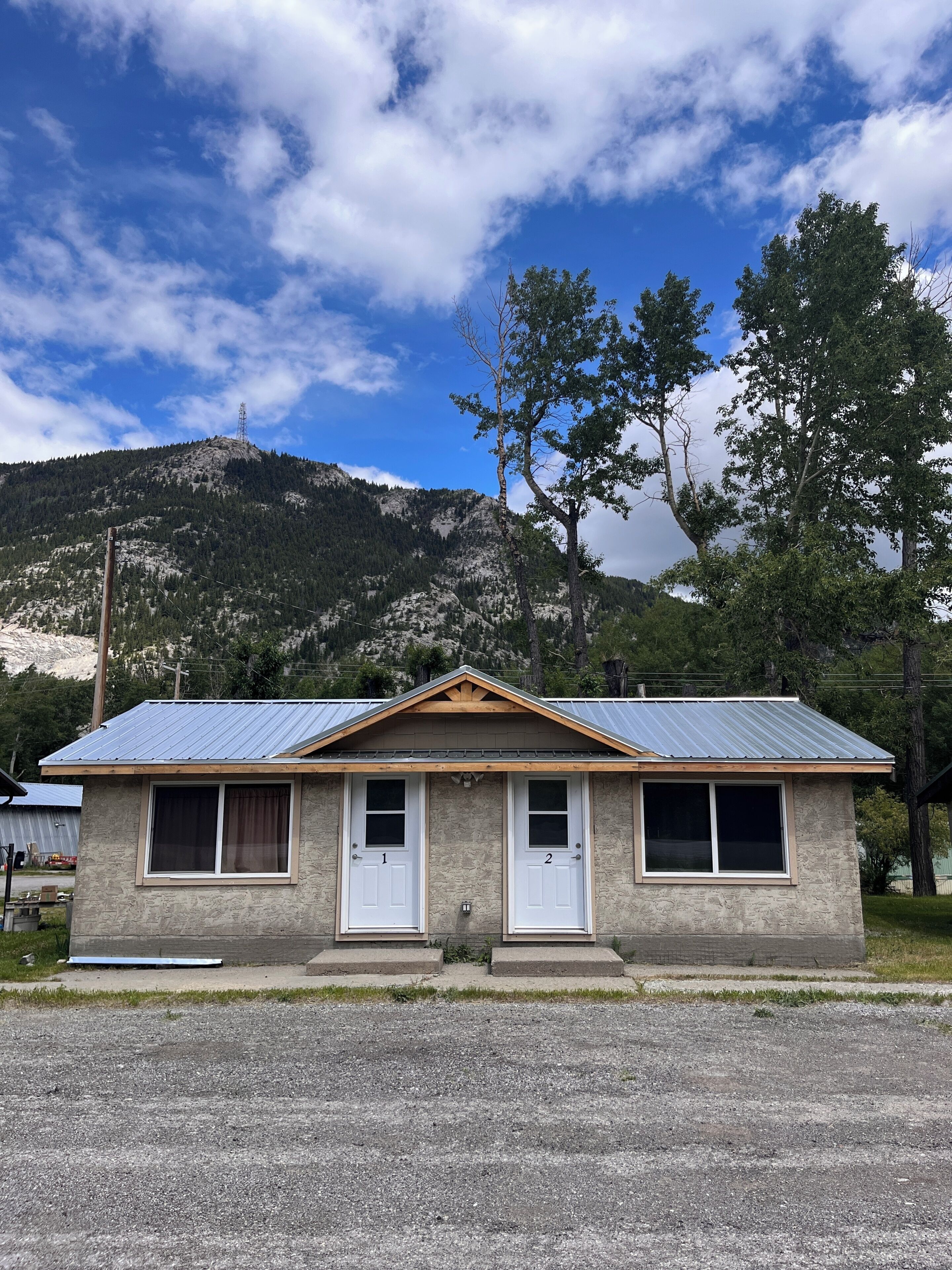 A small building with a metal roof, two doors, and windows. It is located in a rural area with a mountain in the background.