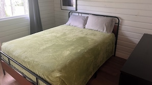 Een enkel bed met een metalen frame, een groene spreidekking en twee kussens. Een ingelijst beeld hangt aan de muur boven het bed.
