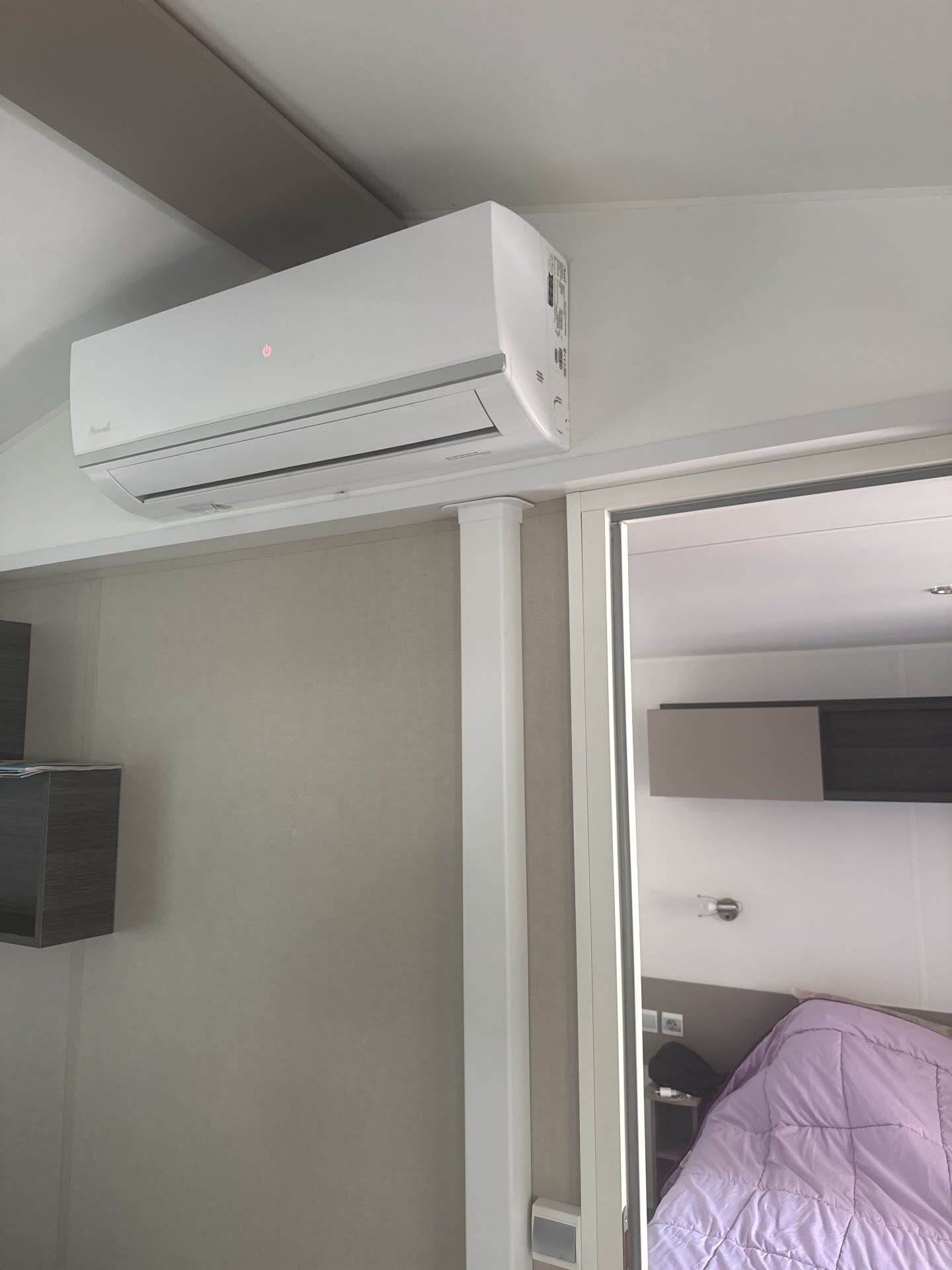 Una habitación con un equipo de aire acondicionado blanco montado en la pared, una puerta que da a otra estancia, y una cama con una manta morada.