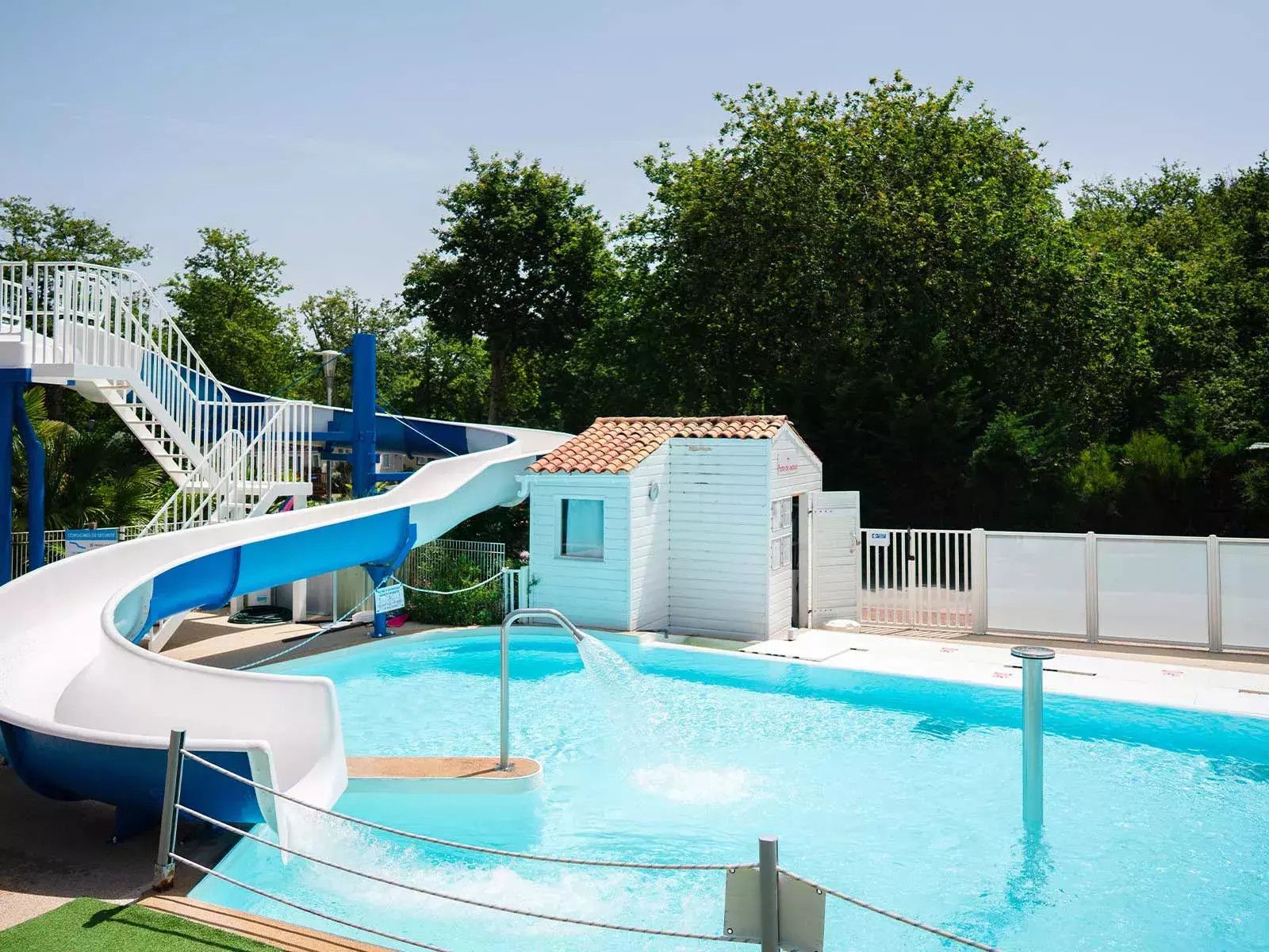Una piscina con un tobogán azul y blanco, un edificio pequeño y un área cercada.
