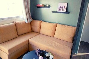 A corner sofa with a small table in front, a window with curtains, and a wall with two shelves holding books and a framed picture. - Mobilhome 2 chambres 2 SDB (Canet-en-Roussillon)