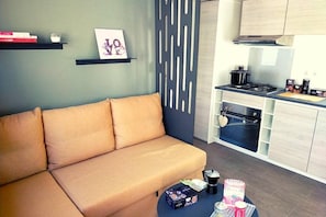 Ein modernes Wohnzimmer mit einer Couch, einem Couchtisch mit Gegenständen, einer Küchenzeile mit Schränken und einem dekorativen Wandbild.