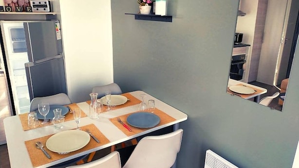 A dining area with a table set for two, a refrigerator, and a small shelf with decorative items. - Mobilhome 2 chambres 2 SDB (Canet-en-Roussillon)
