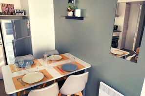 A dining area with a table set for two, a refrigerator, and a small shelf with decorative items. - Mobilhome 2 chambres 2 SDB (Canet-en-Roussillon)