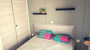 A bedroom with a bed, two pillows, a headboard, a wardrobe, and a shelf with a plant. - Mobilhome 2 chambres 2 SDB (Canet-en-Roussillon)
