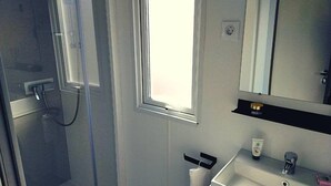 A bathroom with a glass shower enclosure, a sink with a mirror above it, and a window providing natural light. - Mobilhome 2 chambres 2 SDB (Canet-en-Roussillon)