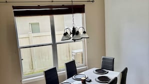 Un coin repas avec une table dressée pour quatre personnes, des chaises, un luminaire suspendu et une fenêtre avec des stores.