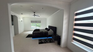 Une chambre à coucher comprenant un lit, une table de chevet, un bureau, un ventilateur de plafond et une fenêtre avec des stores.