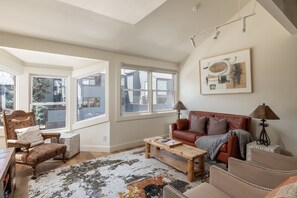 Ein geräumiges Wohnzimmer mit großem Fenster, einem braunen Ledersofa, einem Holztisch und einem gemusterten Teppich.