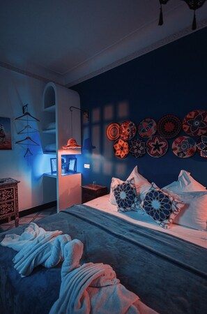 Un dormitorio con cama, almohadas, una lámpara de pared y una estantería con platos decorativos.