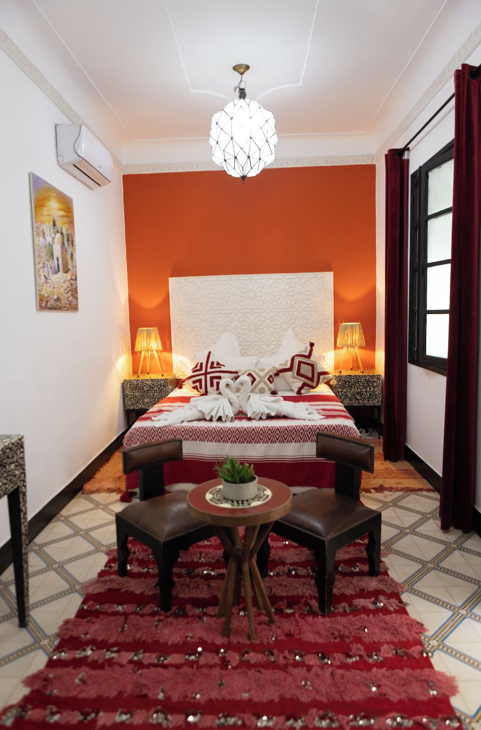 A hotel room with a large bed, two bedside lamps, a chandelier, a red carpet, and a small table with a plant.
