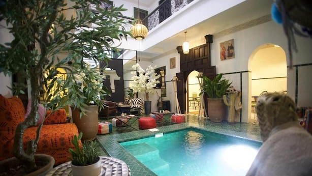 An indoor swimming pool area with a tiled floor, surrounded by potted plants, a seating area with a patterned table, and a staircase leading to an upper level. - RIAD NEW MOON (Marrakech)