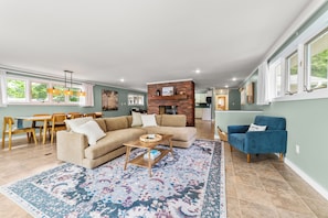A spacious living room with a sectional sofa, a coffee table, and a blue armchair. A dining area with a wooden table and chairs is visible in the background. - Loonar Landing: (Cheboygan)  Lakefront on Long Lake, Sleeps 8 (Cheboygan)