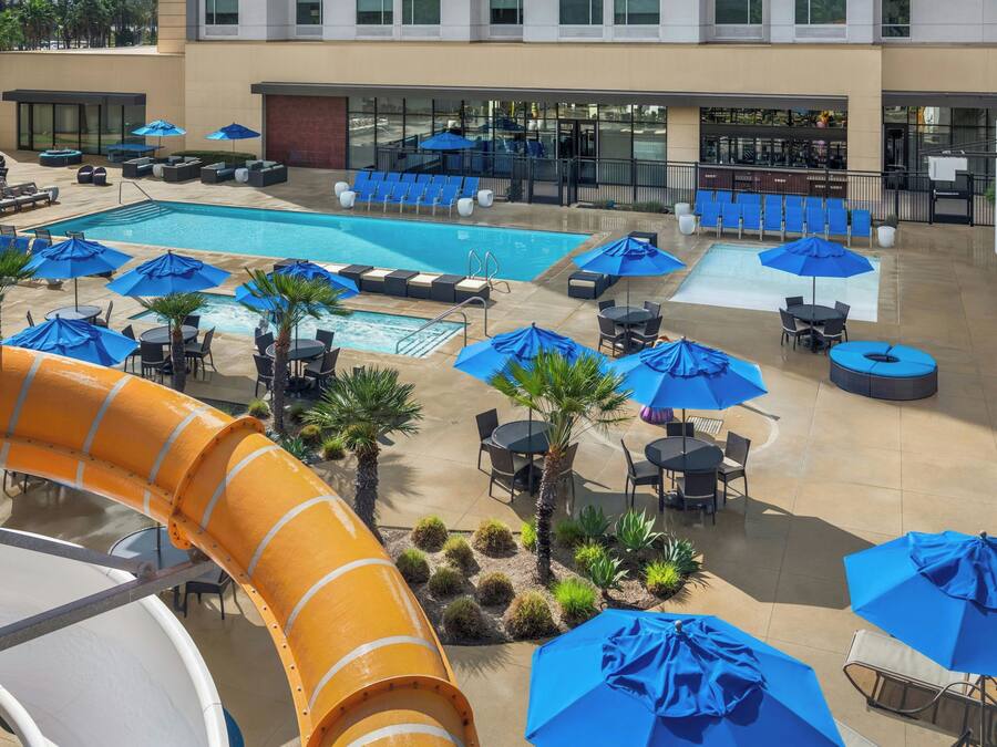A pool area with multiple lounge chairs and umbrellas, a water slide, and a building with large windows in the background.