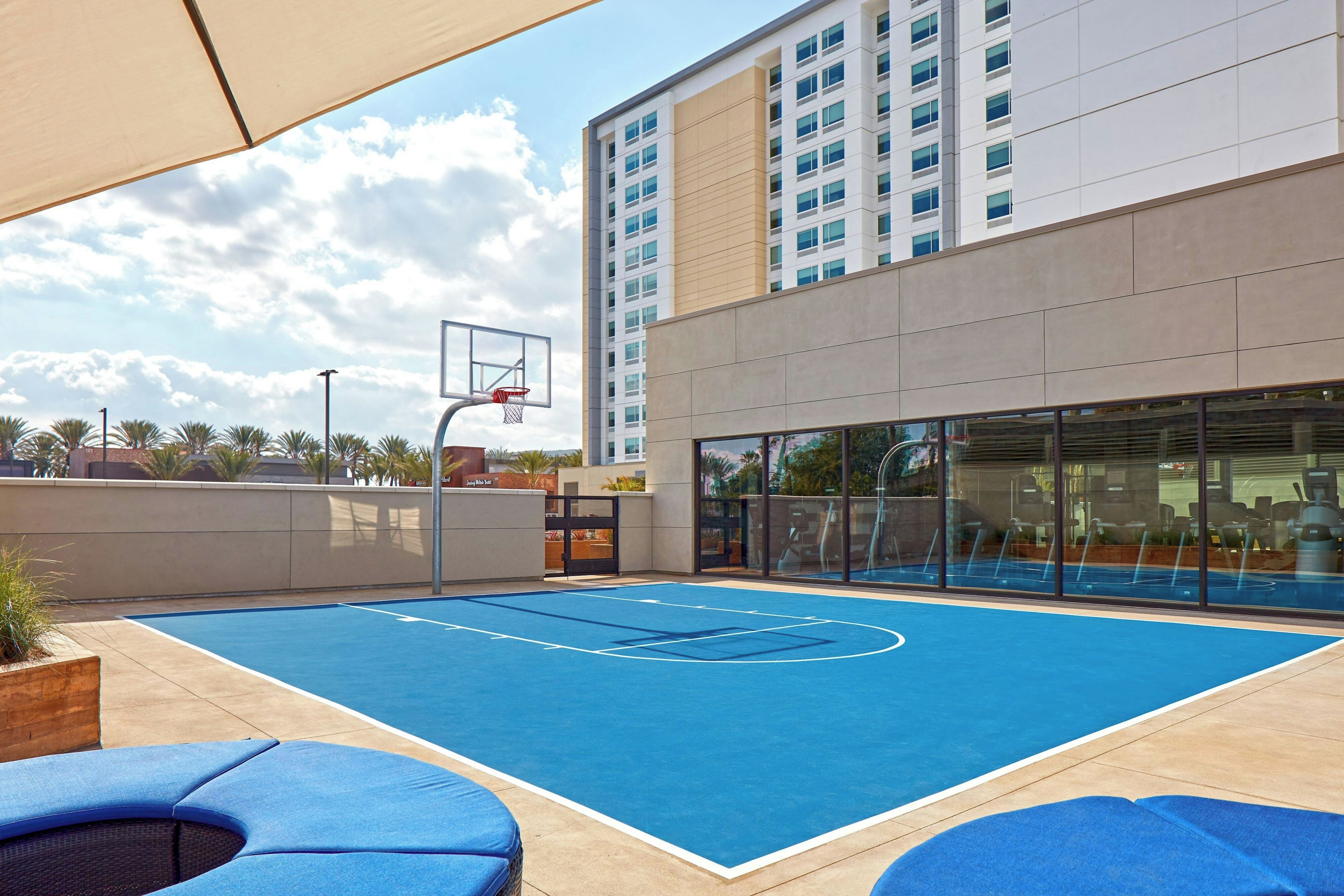 Un terrain de basket en plein air, équipé d’un panier, situé à côté d’un bâtiment doté de grandes fenêtres et d’une porte en verre donnant accès à une salle de sport.