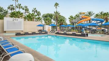 A swimming pool with lounge chairs and umbrellas, surrounded by palm trees and a water slide.