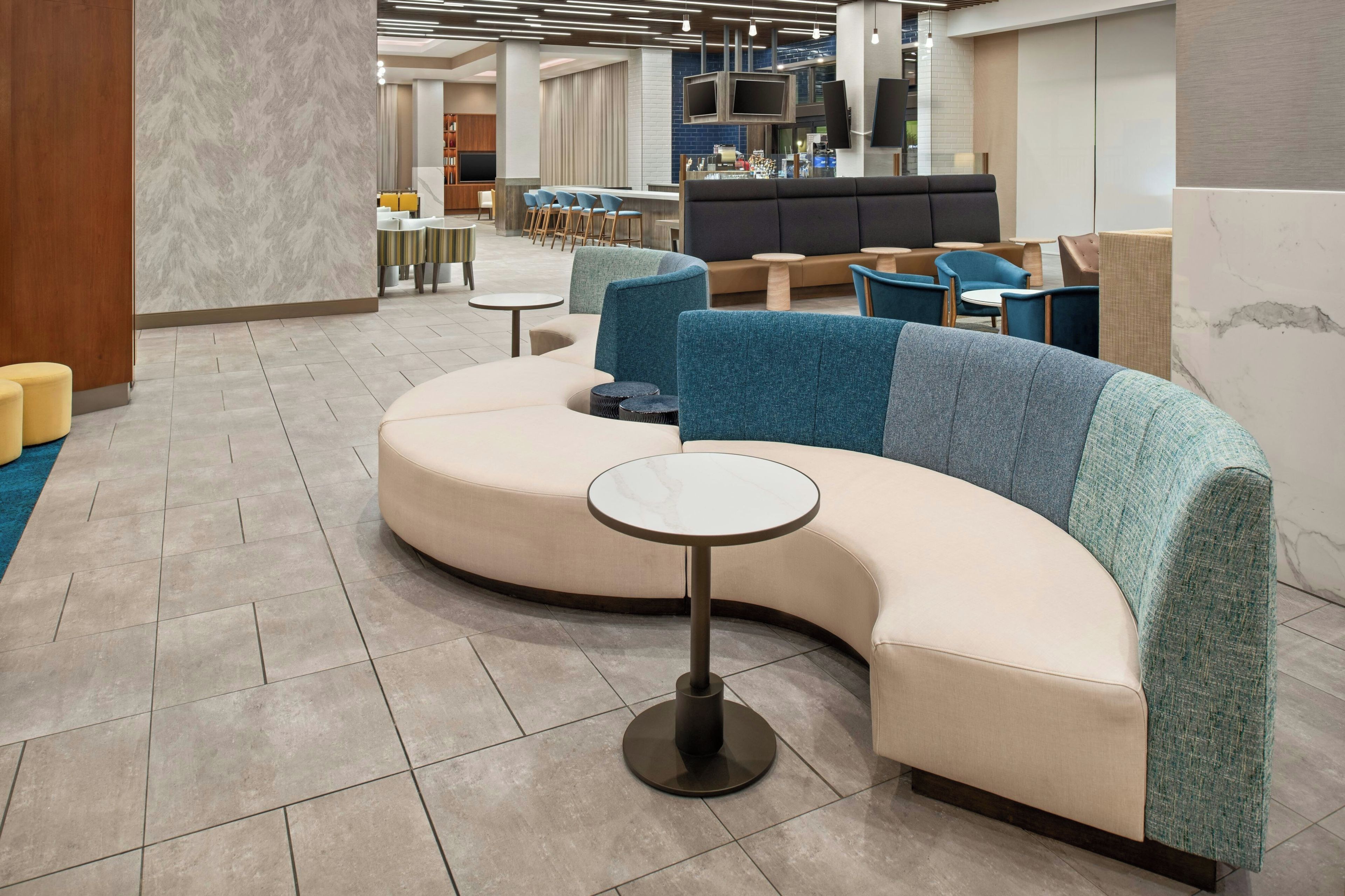 A modern lounge area with a curved sofa, small round tables, and a bar area in the background.