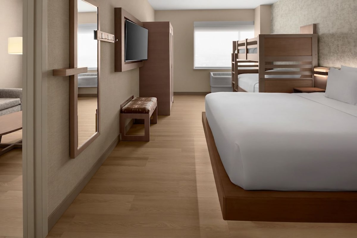 Habitación de hotel con una cama grande, un televisor montado en la pared, un banco y una ventana con persianas.