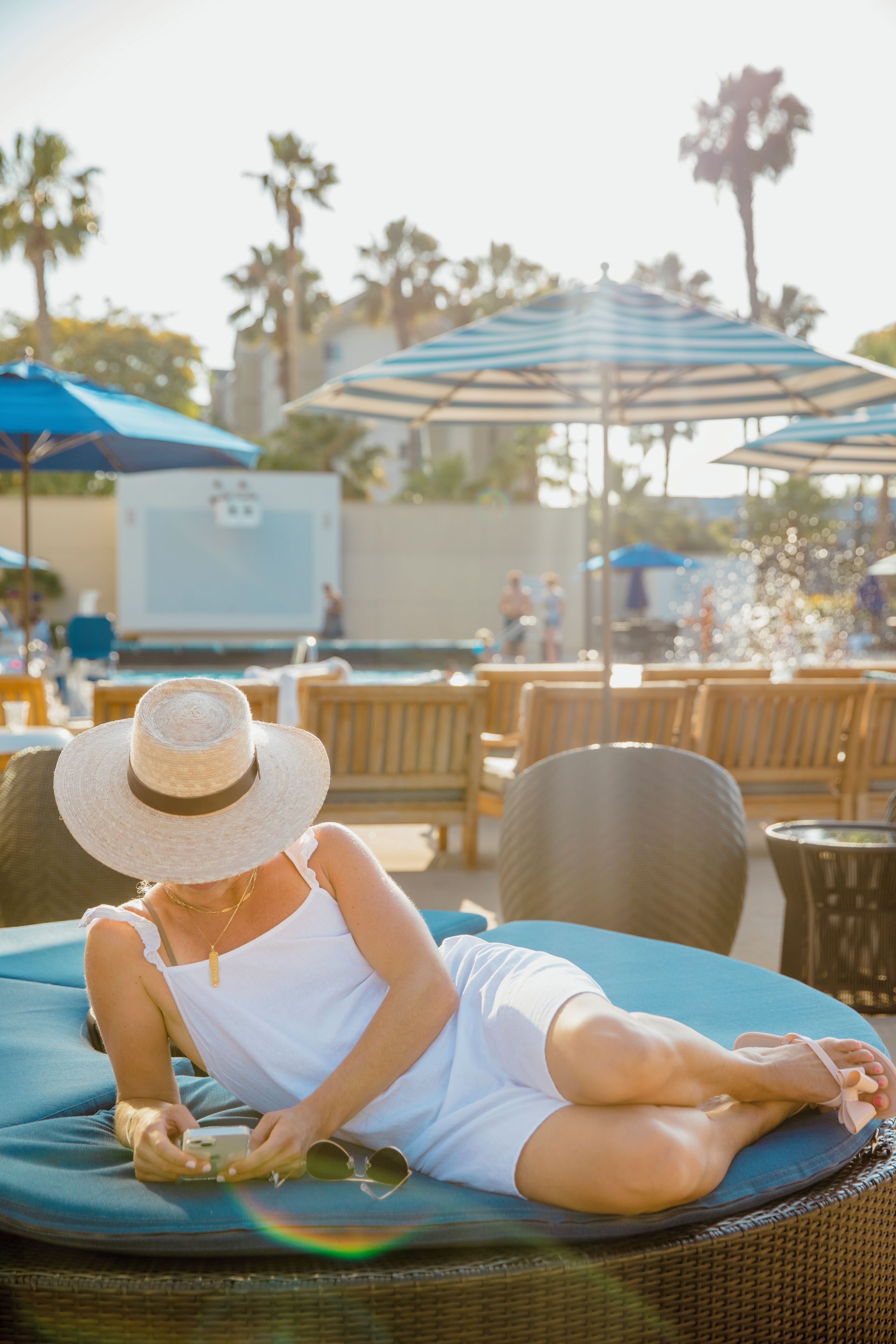 A person sitting on a lounge chair at a poolside area with umbrellas and wooden chairs in the background.