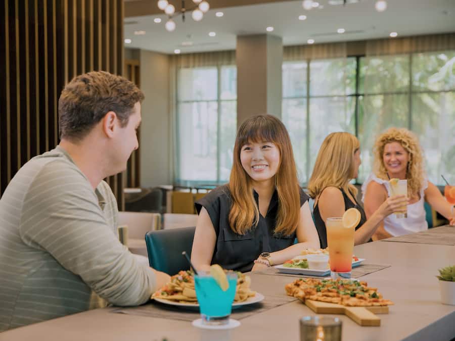 A group of people dining at a restaurant with a modern interior, including a large window and a table with various dishes and drinks.